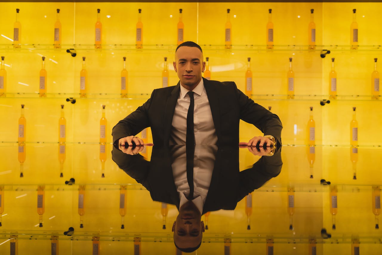 Stylish businessman in suit seated with reflecting surface, surrounded by bottles on a yellow backdrop.