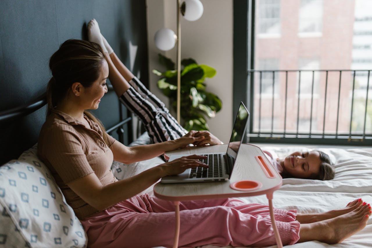 A mother and daughter bonding in a cozy bedroom while working remotely on a laptop.